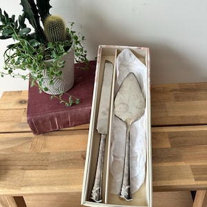 May include: A silver-toned cake serving set consisting of a serrated knife and a triangular server, both featuring ornate floral detailing on the handles. The utensils are presented in a rectangular box lined with white tissue paper.