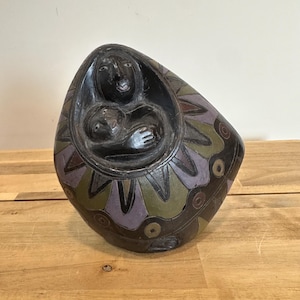 May include: A dark, egg-shaped sculpture depicting a mother and child. The figures are carved in a dark color, with the outer surface decorated with geometric patterns in green, purple, and brown. The sculpture is placed on a wooden surface.