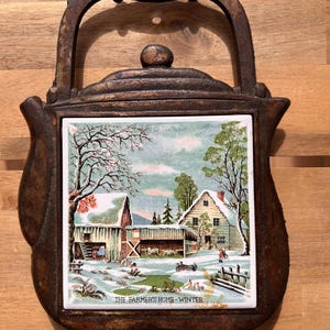Puede incluir: Un salvamanteles de hierro fundido con asa e incrustación de baldosa cerámica. La baldosa presenta una escena invernal con una granja, un granero y el texto "THE FARMER'S HOME - WINTER". El salvamanteles es marrón y tiene un aspecto rústico.