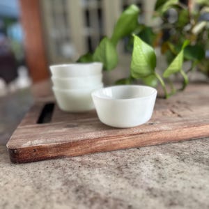 May include: A stack of four small, white, round bowls and one bowl in front, on a wooden cutting board. The bowls are made of a milky white material. Green leaves are in the background.