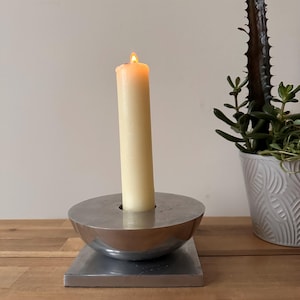 May include: A lit, ivory-colored candle in a modern, silver-toned metal candle holder. The candle holder has a rounded bowl shape on a rectangular base. A potted succulent plant is in the background.