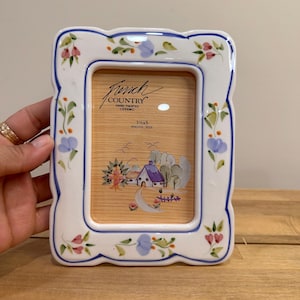 May include: White ceramic photo frame with a scalloped edge, blue trim, and floral accents. The frame has the text "French Country" and is designed for a 3.5 x 5 inch photo. The frame has a painted landscape scene.