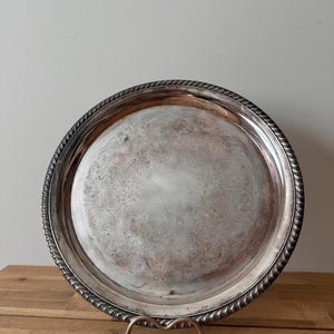 Puede incluir: Una bandeja redonda de color plateado con un borde decorativo en forma de cuerda. La bandeja tiene un intrincado diseño floral grabado en el centro. Se exhibe sobre un soporte de metal dorado, adecuado para servir alimentos o como pieza decorativa.