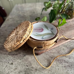 May include: A woven basket with a lid, containing a stack of plates. The plates feature a floral design with pink and green accents. A tan rope handle is attached to the basket. The basket is sitting on a wooden board.
