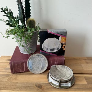 May include: A 7-piece silver-plated coaster set in a box labeled Silver Plated 7pcs Coaster Set. The round coasters feature an embossed pineapple design. One coaster is displayed upright, while the others are stacked in a metal holder.