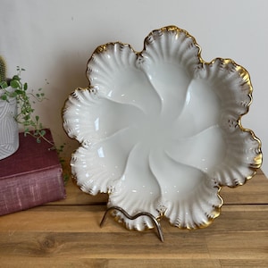 May include: A white ceramic oyster plate with six shell-shaped compartments, featuring a scalloped edge and gold-painted trim. The plate has a glossy finish and a central circular depression, supported by a metal stand for display.