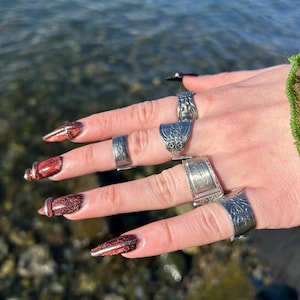 May include: A hand displaying several silver rings of varying designs. The rings include ornate patterns and unique shapes. The nails are painted with a glittery red polish. The background shows water.