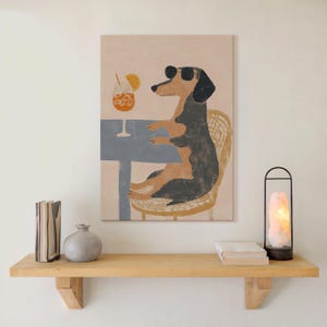 May include: A canvas print depicts a dachshund wearing sunglasses, seated at a table with a cocktail. The dog is in a wicker chair, with a blue table and a drink with an orange slice. The artwork has a neutral color palette.