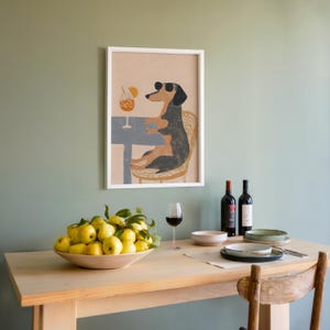 May include: A framed art print depicts a dachshund wearing sunglasses, seated at a table with a cocktail. A wooden table is set with a bowl of lemons, wine glasses, and bottles of wine. The wall is painted a muted green.