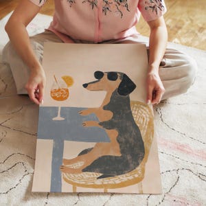 May include: A whimsical illustration of a dachshund wearing sunglasses, seated at a table with a cocktail. The dog is depicted in black and tan, with a blue table and a cocktail glass with ice and an orange slice.