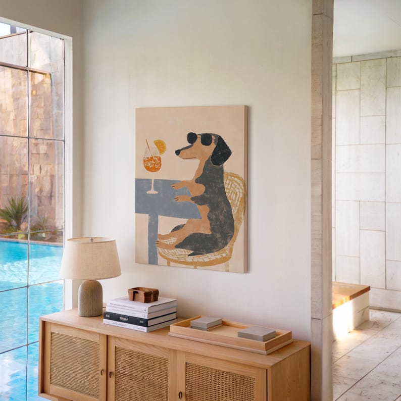 May include: A rectangular canvas art print depicts a dachshund wearing sunglasses, seated at a table with a cocktail. The dog is black and brown, and the table is blue. The background is a light beige.