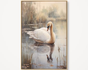 May include: A framed art print depicts a serene lake scene. A white swan with a curved neck floats on the water, its reflection visible. Reeds and grasses line the water's edge, and the background features soft, muted tones of beige and green.