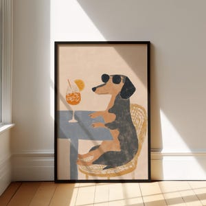 May include: A framed art print featuring a dachshund wearing sunglasses, seated at a table with a cocktail. The dog is depicted in a relaxed pose, with a drink and orange slice. The artwork has a whimsical, illustrative style.