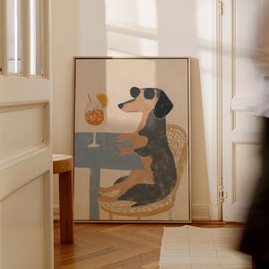 May include: A framed art print featuring a dachshund wearing sunglasses, seated at a table with a cocktail. The dog is depicted in a relaxed pose, seated on a wicker chair. The artwork has a light color palette with a touch of blue.