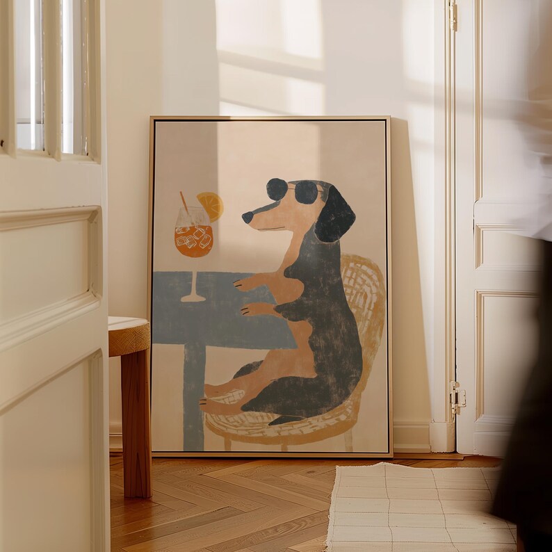 May include: A framed art print featuring a dachshund wearing sunglasses, seated at a table with a cocktail. The dog is depicted in a relaxed pose, seated on a wicker chair. The artwork has a light color palette with a touch of blue.