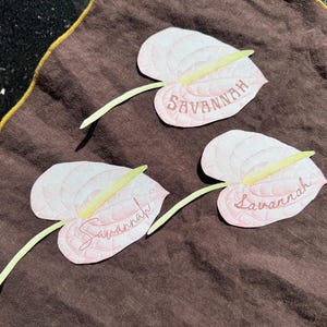 May include: Three light pink paper flower cutouts with the name "Savannah" written on them, each with a yellow stamen and green stem, arranged on a brown cloth with a gold border.