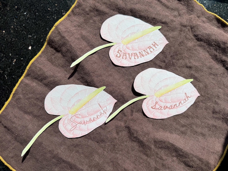 May include: Three light pink paper flower cutouts with the name "Savannah" written on them, each with a yellow stamen and green stem, arranged on a brown cloth with a gold border.