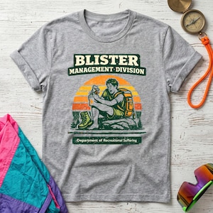 May include: A gray t-shirt with the text "BLISTER MANAGEMENT DIVISION" above an illustration of a hiker treating a blister. The text "Department of Recreational Suffering" is below the illustration. The hiker is wearing a backpack and hiking boots.