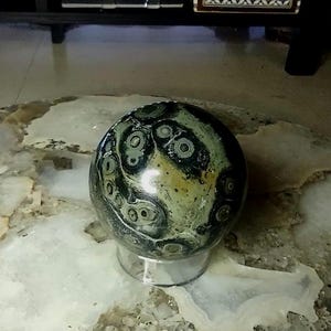 May include: A polished orb of green and black rhyolite stone featuring distinct circular patterns and eye-like inclusions. The spherical stone rests on a clear, circular acrylic stand, showcasing a smooth, reflective surface with earthy tones.