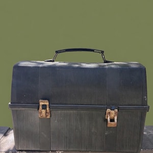 Puede incluir: Una fiambrera negra de estilo vintage con textura acanalada. Cuenta con un asa negra y dos cierres met&aacute;licos oxidados. La fiambrera rectangular est&aacute; sobre una superficie de hormig&oacute;n contra un fondo verde.