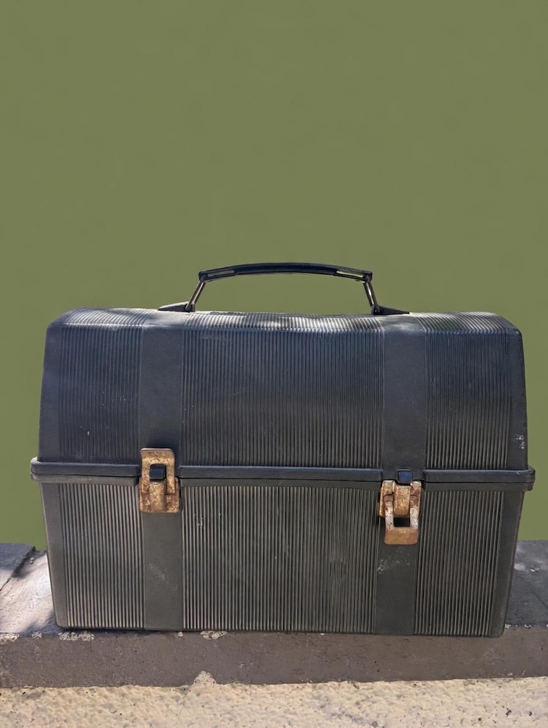 Puede incluir: Una fiambrera negra de estilo vintage con textura acanalada. Cuenta con un asa negra y dos cierres met&aacute;licos oxidados. La fiambrera rectangular est&aacute; sobre una superficie de hormig&oacute;n contra un fondo verde.