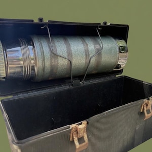 Puede incluir: Termo verde y plateado vintage dentro de una fiambrera de pl&aacute;stico negro. El termo se sujeta con un soporte met&aacute;lico. La fiambrera tiene una textura acanalada y cierres met&aacute;licos oxidados. El fondo es verde oliva liso.