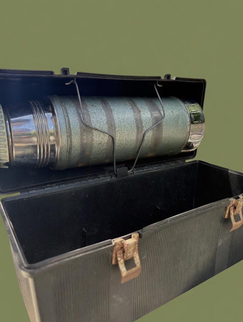 Puede incluir: Termo verde y plateado vintage dentro de una fiambrera de pl&aacute;stico negro. El termo se sujeta con un soporte met&aacute;lico. La fiambrera tiene una textura acanalada y cierres met&aacute;licos oxidados. El fondo es verde oliva liso.