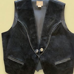 May include: Black suede vest with a V-neck design and two decorative buttons. Features two front pockets and a Wrangler label. A classic western-style garment.