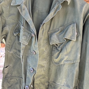 Puede incluir: Chaqueta verde oliva de estilo militar con múltiples bolsillos y cierres de botón. La chaqueta tiene cuello y parece estar hecha de una tela duradera. La prenda muestra signos de desgaste, lo que se suma a su estética vintage.