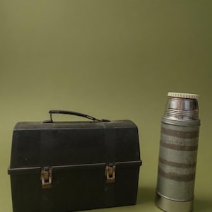Puede incluir: Una fiambrera negra vintage con asa y cierres met&aacute;licos, junto a un termo a juego. El termo tiene tapa plateada y un dise&ntilde;o a rayas verdes y plateadas. Ambos art&iacute;culos est&aacute;n sobre un fondo verde.