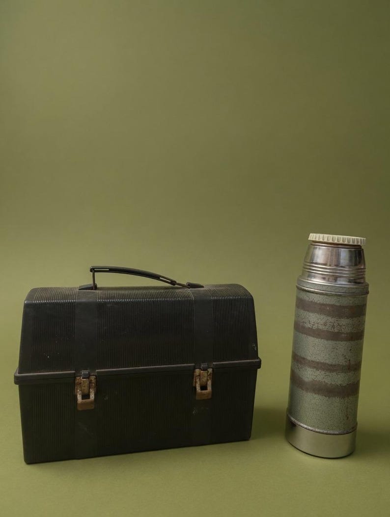 Puede incluir: Una fiambrera negra vintage con asa y cierres met&aacute;licos, junto a un termo a juego. El termo tiene tapa plateada y un dise&ntilde;o a rayas verdes y plateadas. Ambos art&iacute;culos est&aacute;n sobre un fondo verde.