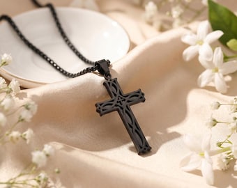 Pode incluir: Um colar com pingente de cruz preta com um design intrincado. A cruz é suspensa por uma corrente preta combinando. O colar repousa sobre um tecido de cetim creme, rodeado por flores brancas.