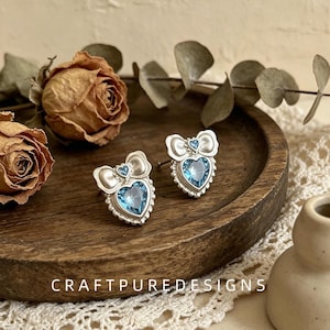 May include: A pair of silver-toned stud earrings featuring a central heart-shaped light blue crystal, a smaller blue crystal accent, and a bow-shaped top with a beaded border. The text CRAFTPUREDESIGNS is visible at the bottom of the image.