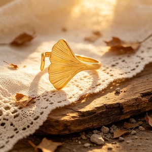 May include: A gold-colored ring with a ginkgo leaf design. The ring is open and adjustable, with the leaf shape prominently displayed. The ring is set against a rustic wooden surface with a lace cloth and dried leaves.