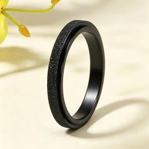 May include: A black ring with a textured, glittery band. The ring is made of a dark, possibly metallic material. The ring is a simple, elegant design.