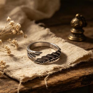 Op de afbeelding: Een zilverkleurige ring met een veermotief. De ring heeft gedetailleerde veermotieven aan beide zijden, waardoor een open, verstelbaar ontwerp ontstaat. De ring wordt gepresenteerd op een beige doek, met een vage achtergrond.