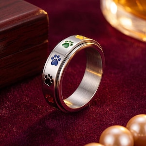 Puede incluir: Un anillo giratorio plateado con huellas de patas coloridas. El anillo presenta una banda exterior giratoria con huellas de patas en rojo, azul, verde y amarillo. El anillo está sobre una superficie de terciopelo rojo.