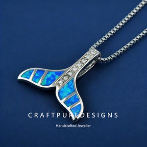 May include: A silver pendant necklace featuring a whale tail design. The tail is inlaid with blue and green opal stones and accented with clear crystals. The necklace hangs on a silver chain against a blue background. The text "CRAFTPUREDESIGNS" and "Handcrafted Jeweller" are visible.