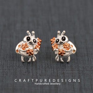 Può includere: Un paio di orecchini a lobo a forma di coccinella color argento. Ogni orecchino presenta una coccinella sorridente che tiene in mano un grappolo di fiori color rame. Gli orecchini hanno occhi neri e antenne.