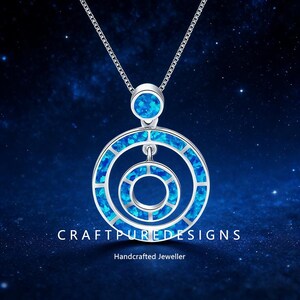 Puede incluir: Un collar de plata con un colgante con un diseño circular con incrustaciones de ópalo azul. El colgante tiene tres círculos concéntricos y un pequeño ópalo redondo en la parte superior. El texto "CRAFTPUREDESIGNS" y "Handcrafted Jeweller" están en la parte inferior.