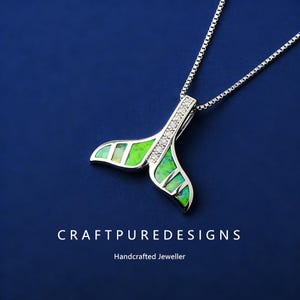 May include: A silver necklace with a pendant shaped like a whale tail. The tail is filled with green and opal-like material and accented with small, clear stones. The necklace is on a blue background. Text on the image reads "CRAFTPUREDESIGNS" and "Handcrafted Jeweller."