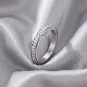 May include: A silver ring with a band of small, clear gemstones. The ring is set against a backdrop of soft, gray fabric, creating a luxurious and elegant feel. The gemstones sparkle, catching the light.