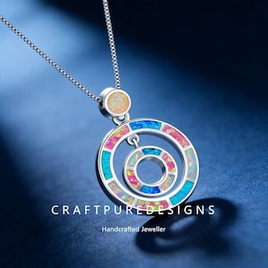 Puede incluir: Un collar de plata con un colgante con dos círculos concéntricos con incrustaciones de piedras de ópalo de colores. El collar tiene una cadena delicada y el texto "CRAFTPUREDESIGNS" y "Handcrafted Jeweller" son visibles.