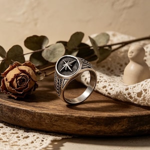May include: A silver ring featuring a compass rose design. The ring has a circular face with a raised starburst emblem. The band features detailed geometric patterns. The ring is displayed on a wooden surface with a dried rose and decorative elements.
