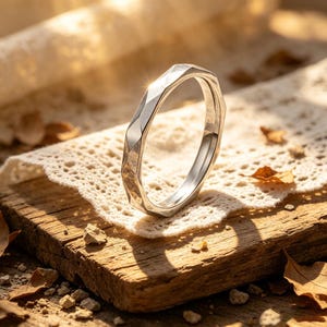 Könnte beinhalten: Ein silberner Ring mit einem geometrischen, facettierten Design. Der Ring befindet sich vor einem Hintergrund aus Holz, Spitze und getrockneten Blättern, was eine rustikale und elegante Ästhetik erzeugt. Die Oberfläche des Rings reflektiert das Licht.