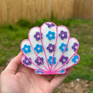 May include: A white seashell-shaped object with a textured surface, outlined in bright pink glitter. The shell is decorated with small, glittery flower shapes in alternating shades of purple and light blue, arranged in a radial pattern.