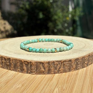 Könnte beinhalten: Ein elastisches Armband bestehend aus kleinen, runden, hellgrünen, türkis-farbenen Perlen, die mit vier glatten, polierten goldfarbenen Metall-Zwischenperlen symmetrisch um das kreisförmige Band angeordnet sind.