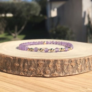 Pode incluir: Uma pulseira delicada com pequenas contas redondas de cor violeta claro intercaladas com contas metálicas douradas. A pulseira é exibida em uma fatia de madeira natural, mostrando sua elegância sutil. O fundo está desfocado, destacando a joia.