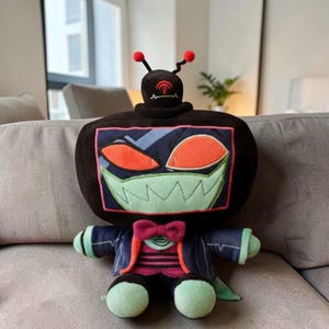 May include: A plush toy shaped like a retro television set with a menacing face. The toy has a black body, red eyes, and a green, jagged-tooth grin. It wears a blue jacket with a pink bow tie and green accents.
