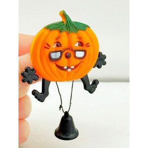 May include: An orange pumpkin-shaped ornament with a green stem, black limbs, and white-rimmed glasses. The pumpkin has a smiling face with two visible teeth. A small black bell hangs from the bottom of the pumpkin by a thin black string.